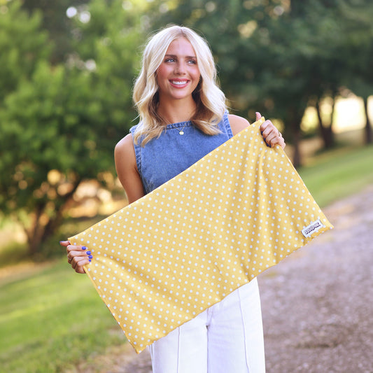 A woman standing outdoors on a gravel path holding up a yellow fabric item with small white polka dots. She is wearing a blue sleeveless top and white pants, with trees and grass in the background.