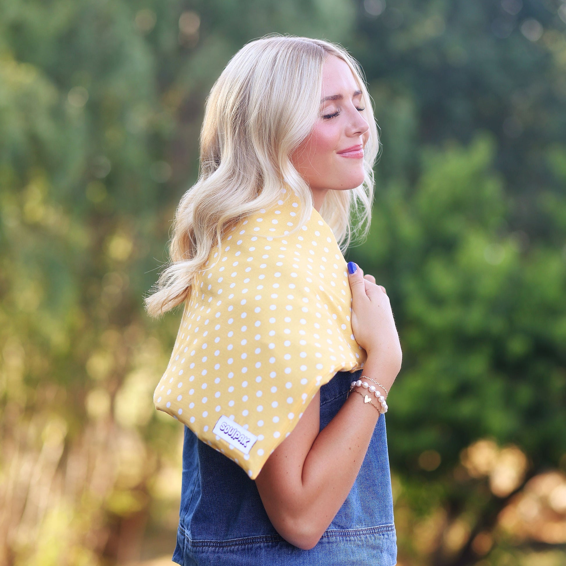 A woman outdoors wearing a yellow and white polka dot pillow around her neck, with a blurred green forest background.