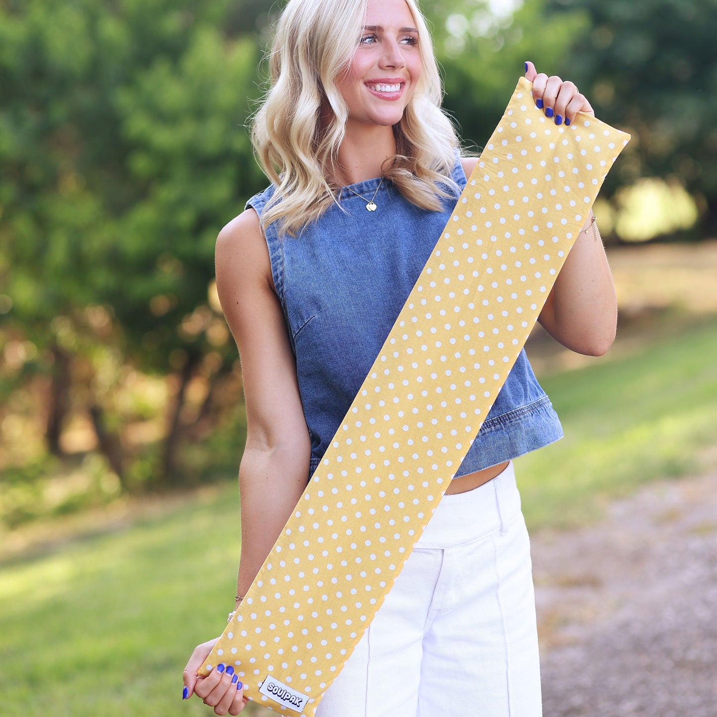 A woman standing outdoors in a park holding a yellow yoga mat with white polka dots.