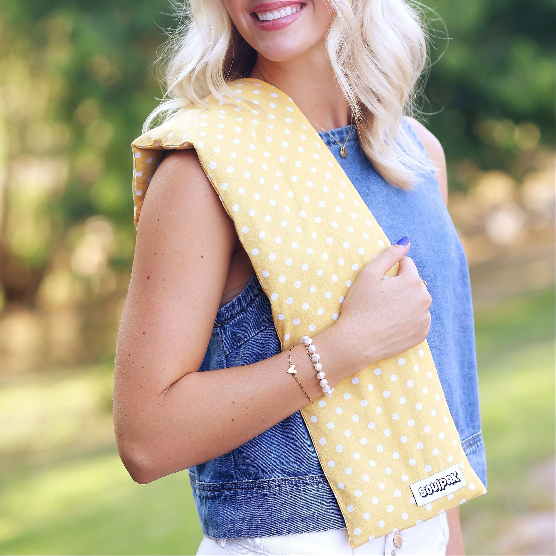 A woman outdoors wearing a yellow and white polka dot baby carrier wrap around her shoulder, paired with a denim vest and white pants.