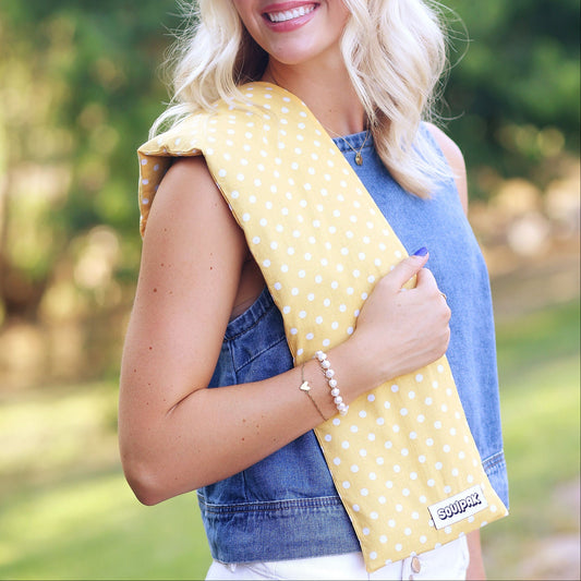 A woman outdoors wearing a yellow and white polka dot baby carrier wrap around her shoulder, paired with a denim vest and white pants.